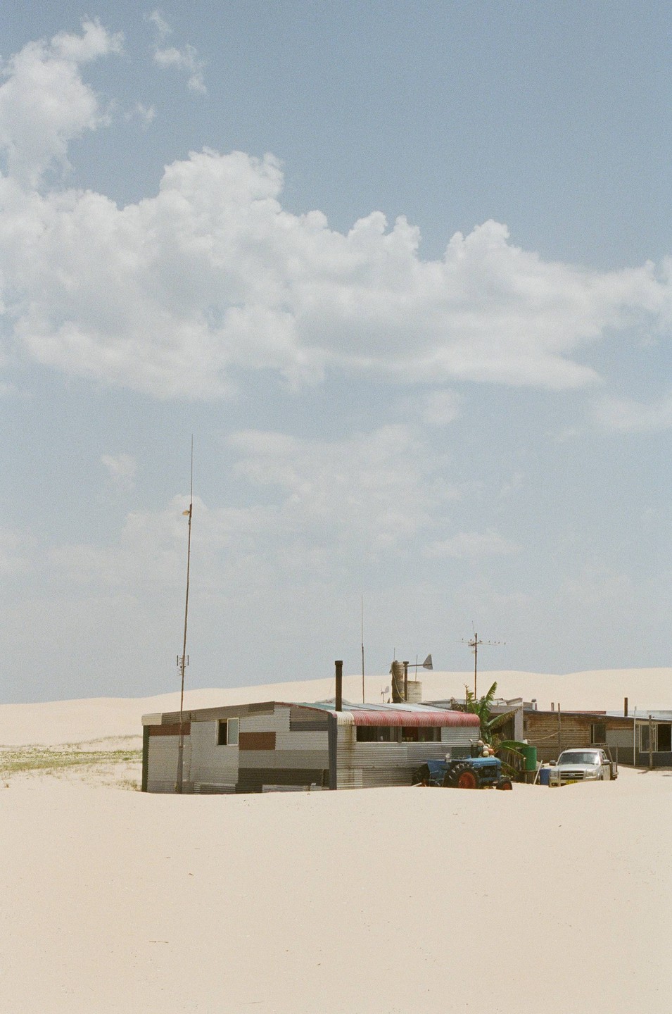A small settlement of corrugated metal buildings is nestled among sand dunes under a cloudy sky.