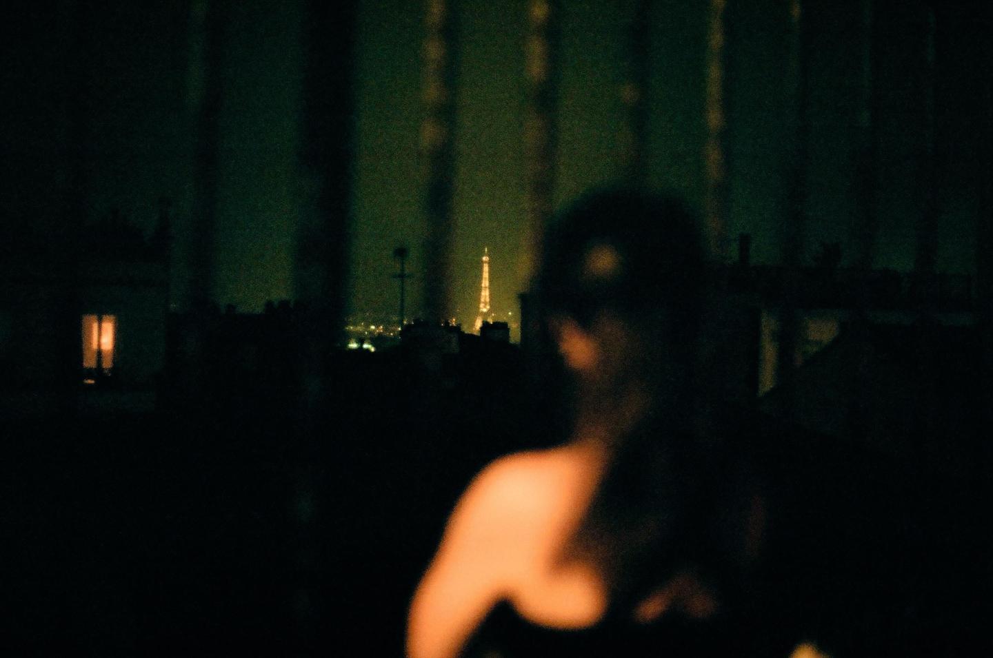 A blurry figure stands in the foreground with the illuminated Eiffel Tower visible in the distance at night.