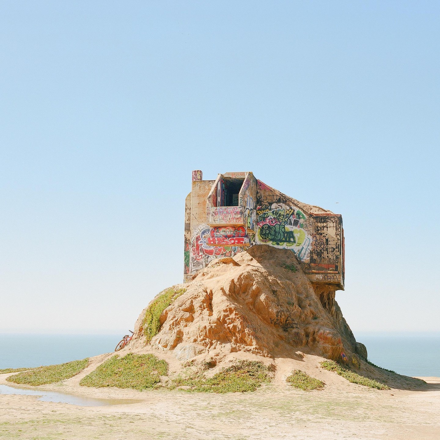 A graffiti-covered concrete structure sits atop a sandy hill overlooking the ocean under a clear sky.