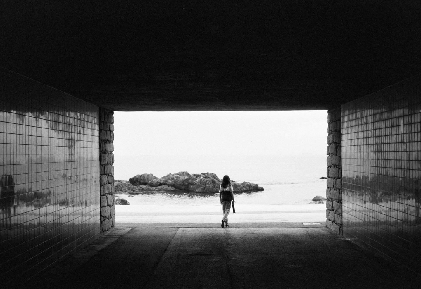 A woman walks out of a tunnel towards a rocky beach and the ocean.