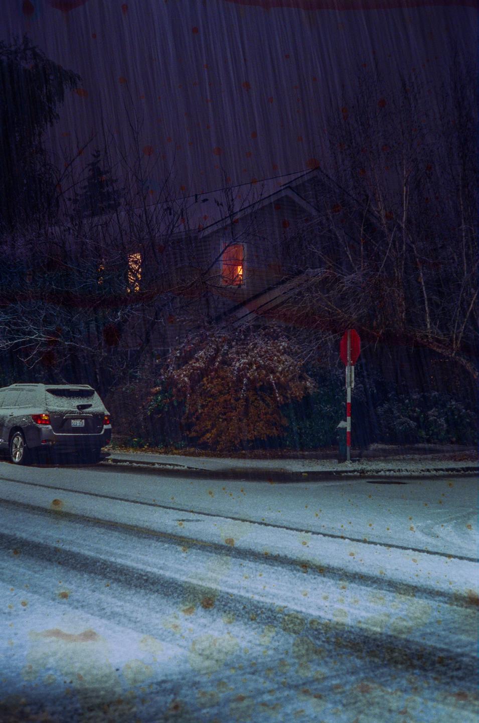 A car is parked on a snowy street at night, with a house and trees in the background.