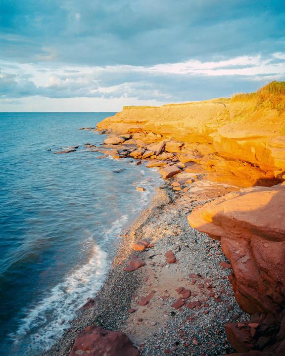 The ocean meets a rocky beach and a cliff under a cloudy sky.