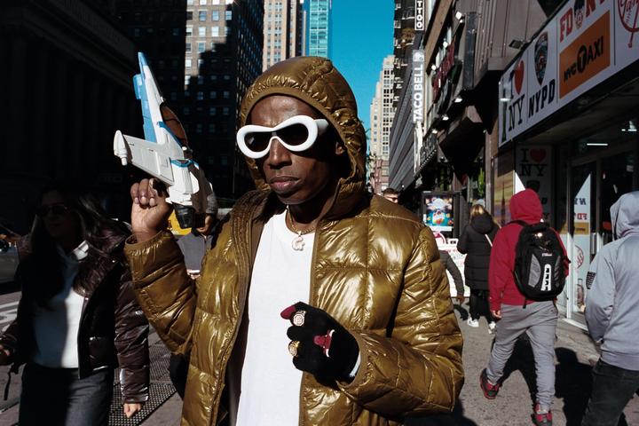 A man in a shiny gold puffer jacket and white sunglasses holds a toy spaceship on a city street.