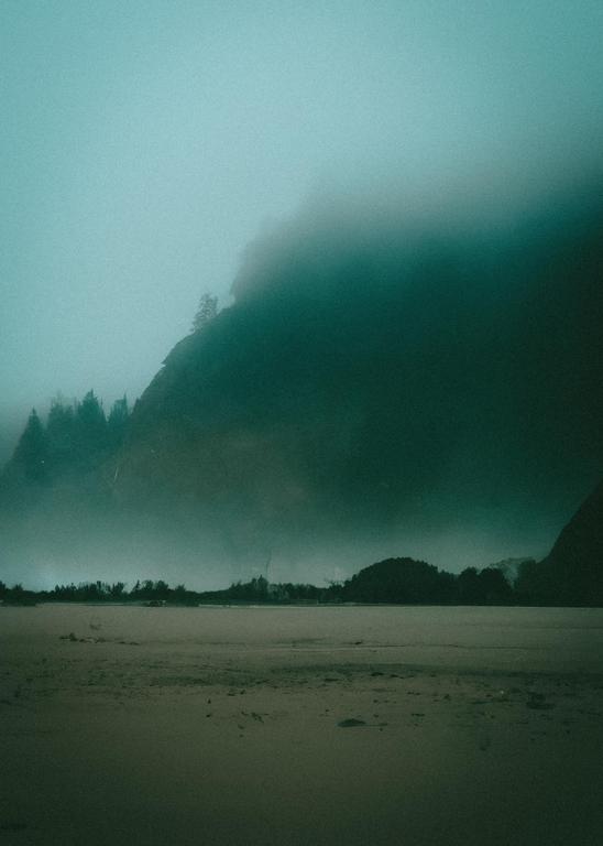 A sandy beach is visible in the foreground, with a body of water and a forested mountain shrouded in fog in the background.