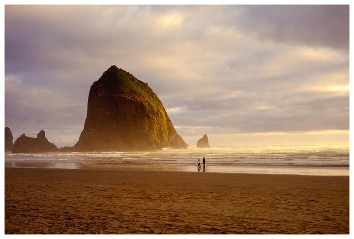 Two people walk on a beach with a large rock formation in the background under a cloudy sky.