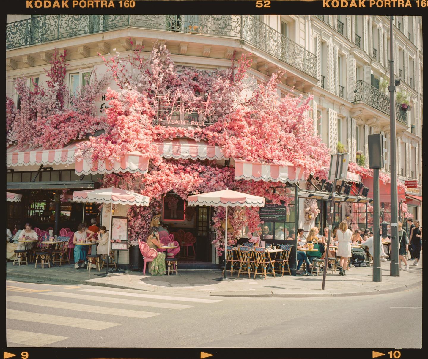 A cafe with pink awnings and pink flowers covering its facade has people seated at tables on the sidewalk.