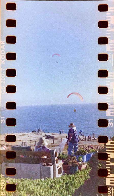 Two people watch paragliders over the ocean from a grassy cliffside.