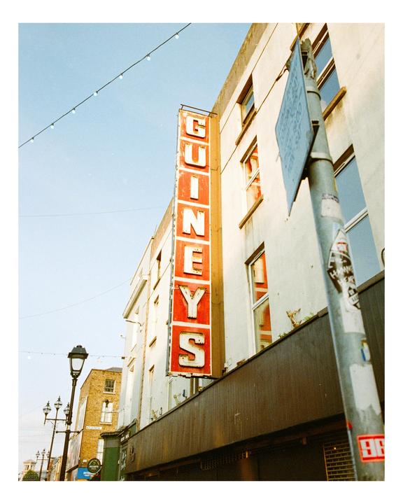 A tall red and white sign for Guineys is attached to the side of a white building on a street in Dublin.