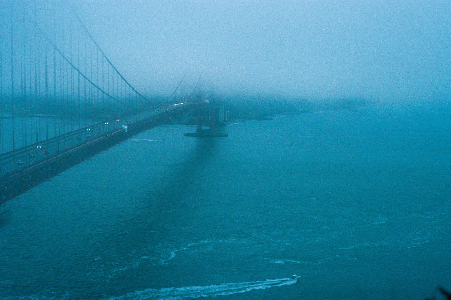 The Golden Gate Bridge is shrouded in fog, with a boat leaving a wake in the water below.