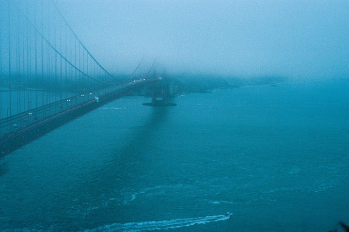 The Golden Gate Bridge is shrouded in fog, with a boat leaving a wake in the water below.