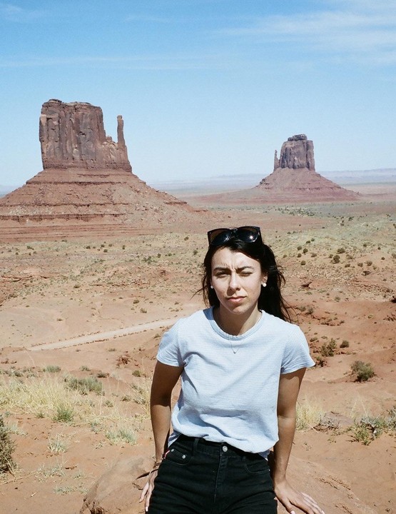 A woman sits on a rock in a desert landscape with large rock formations in the background.