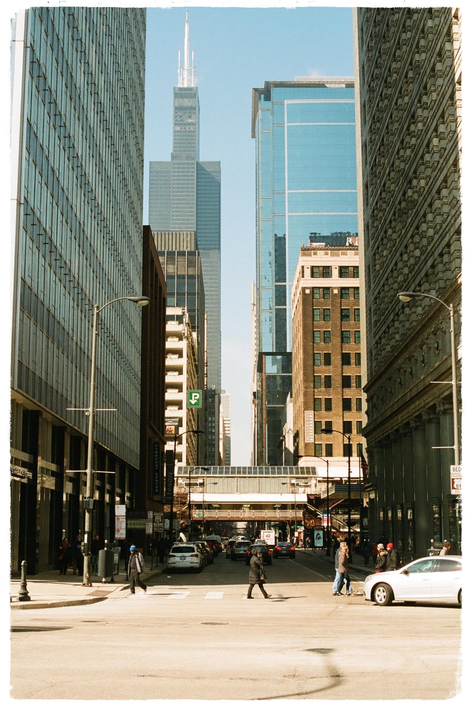 A street in a city is lined with tall buildings and cars, with people crossing the street and a skyscraper visible in the distance.