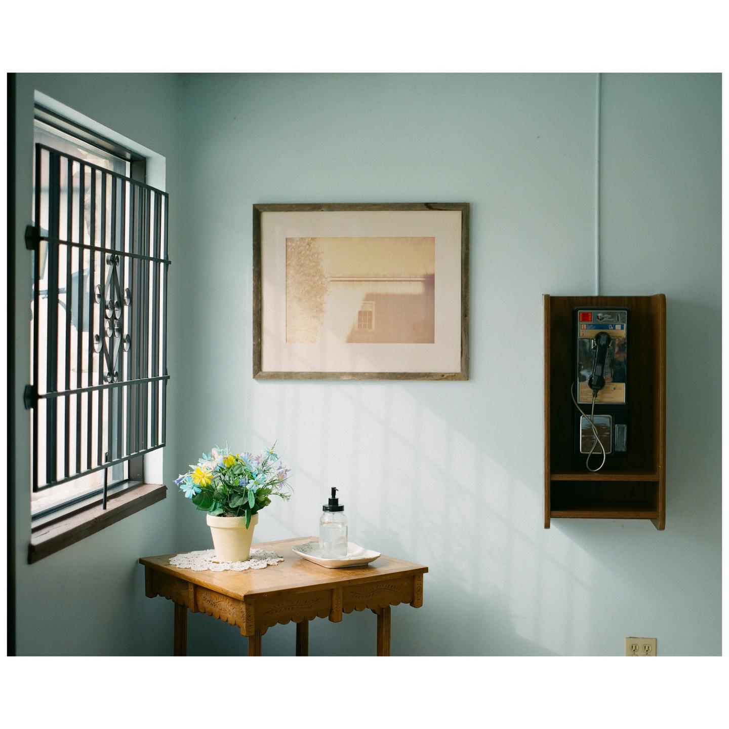 A small wooden table with a potted plant and a bottle sits next to a barred window, with a framed picture and a payphone on the wall.