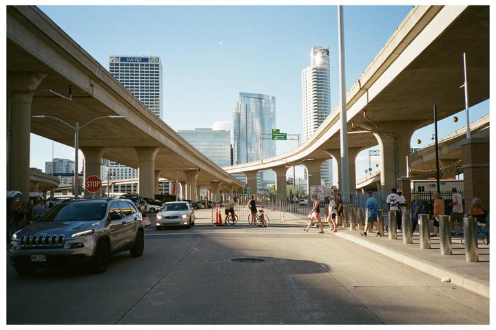 Cars drive on a city street under elevated highways, with people walking on the sidewalk and tall buildings in the background.