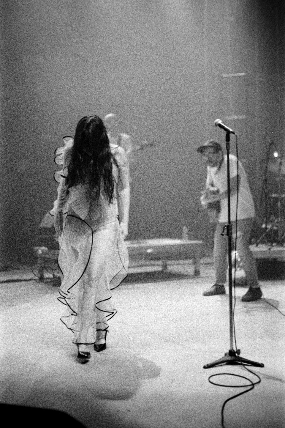 A woman in a ruffled dress walks away from the camera on a stage with a microphone stand in the foreground and a man in the background.