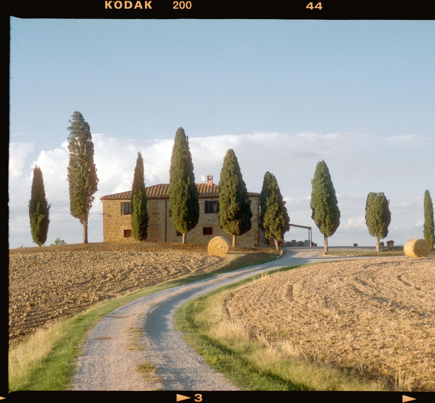 A winding dirt road leads to a stone house surrounded by cypress trees and hay bales in a field.