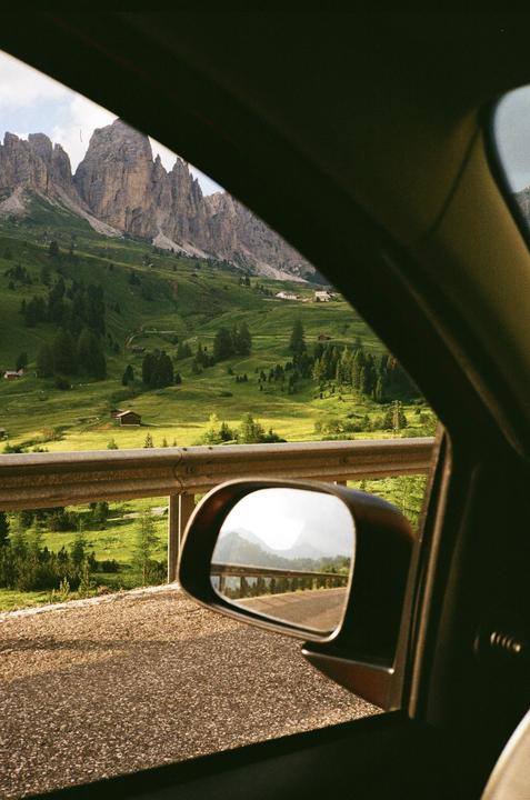 A mountain landscape with green hills and scattered houses is seen from inside a car, with the road and a guardrail reflected in the side mirror.