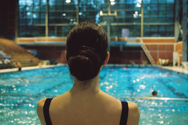 A woman with her hair in a bun looks out over a swimming pool with several people in the water.