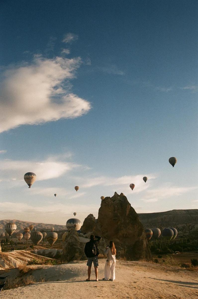 A couple stands on a hill overlooking a valley filled with hot air balloons and rock formations under a blue sky with clouds.