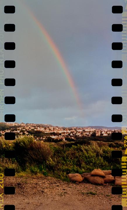 A rainbow arches over a town nestled on a hillside, with green foliage in the foreground.