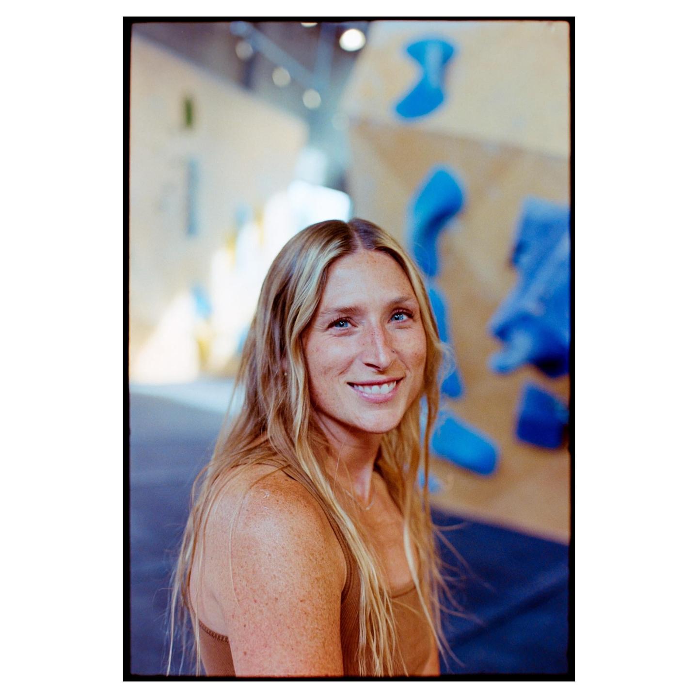 A woman with long blonde hair and freckles smiles at the camera in a climbing gym.