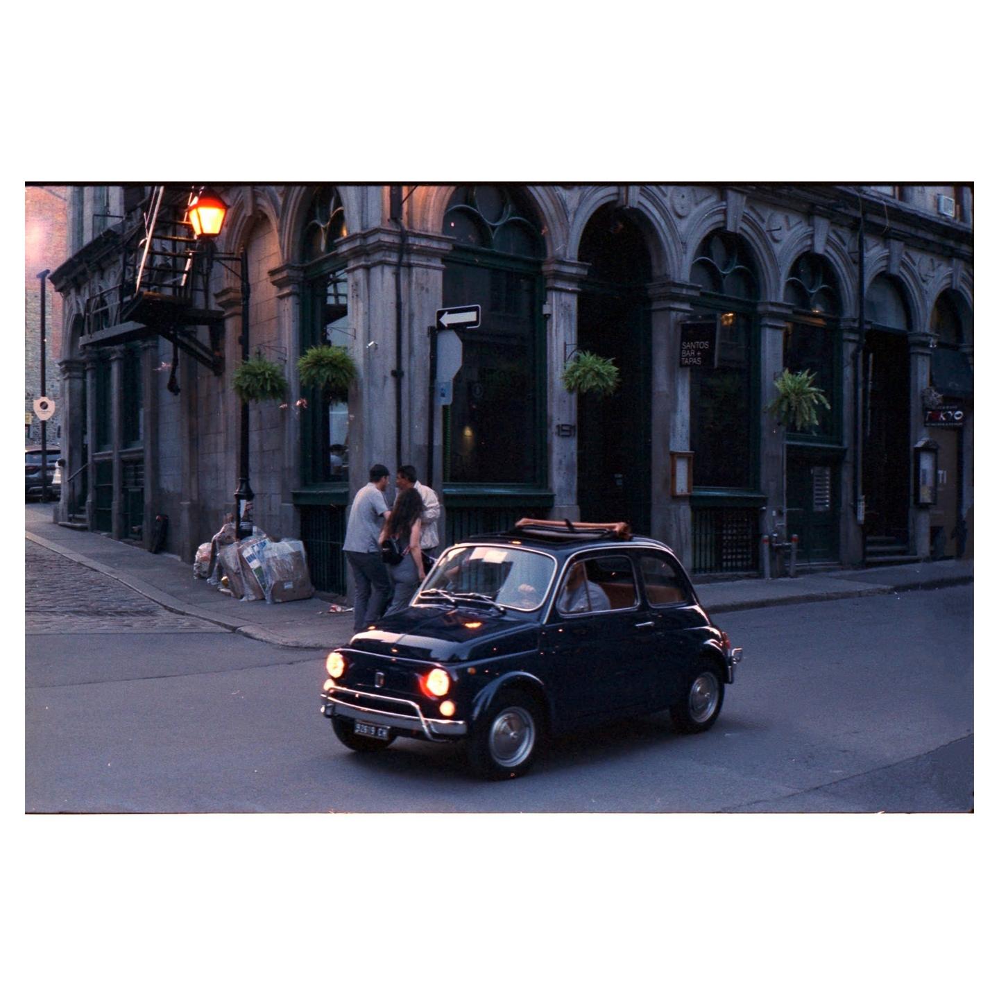 A small dark car with its headlights on drives past a building with arched windows and a street light.