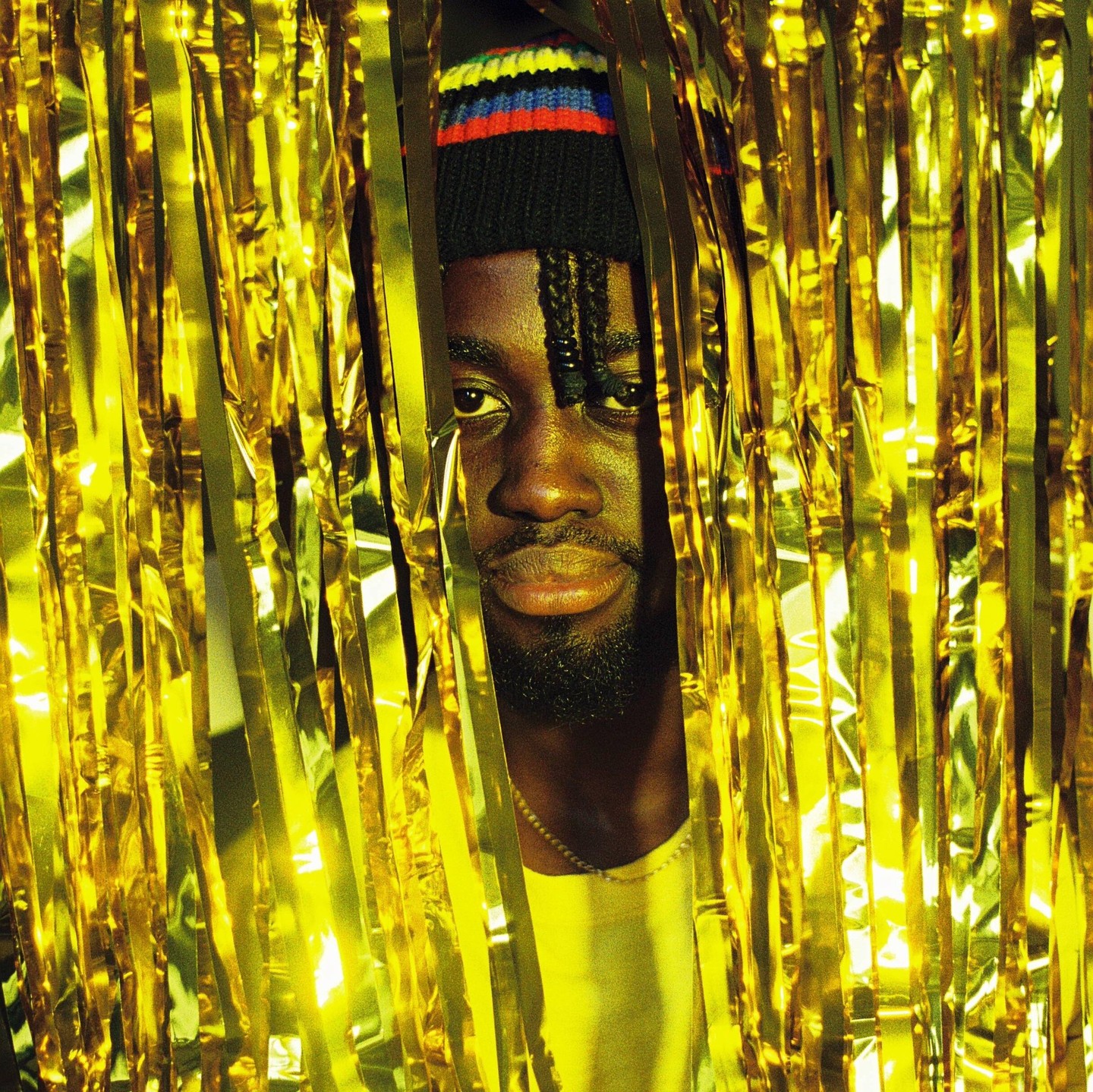 A man with a beard and a colorful beanie peeks through a shimmering gold curtain.