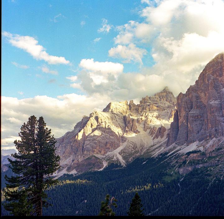 A large mountain range with a forest at its base is illuminated by the sun under a cloudy sky.