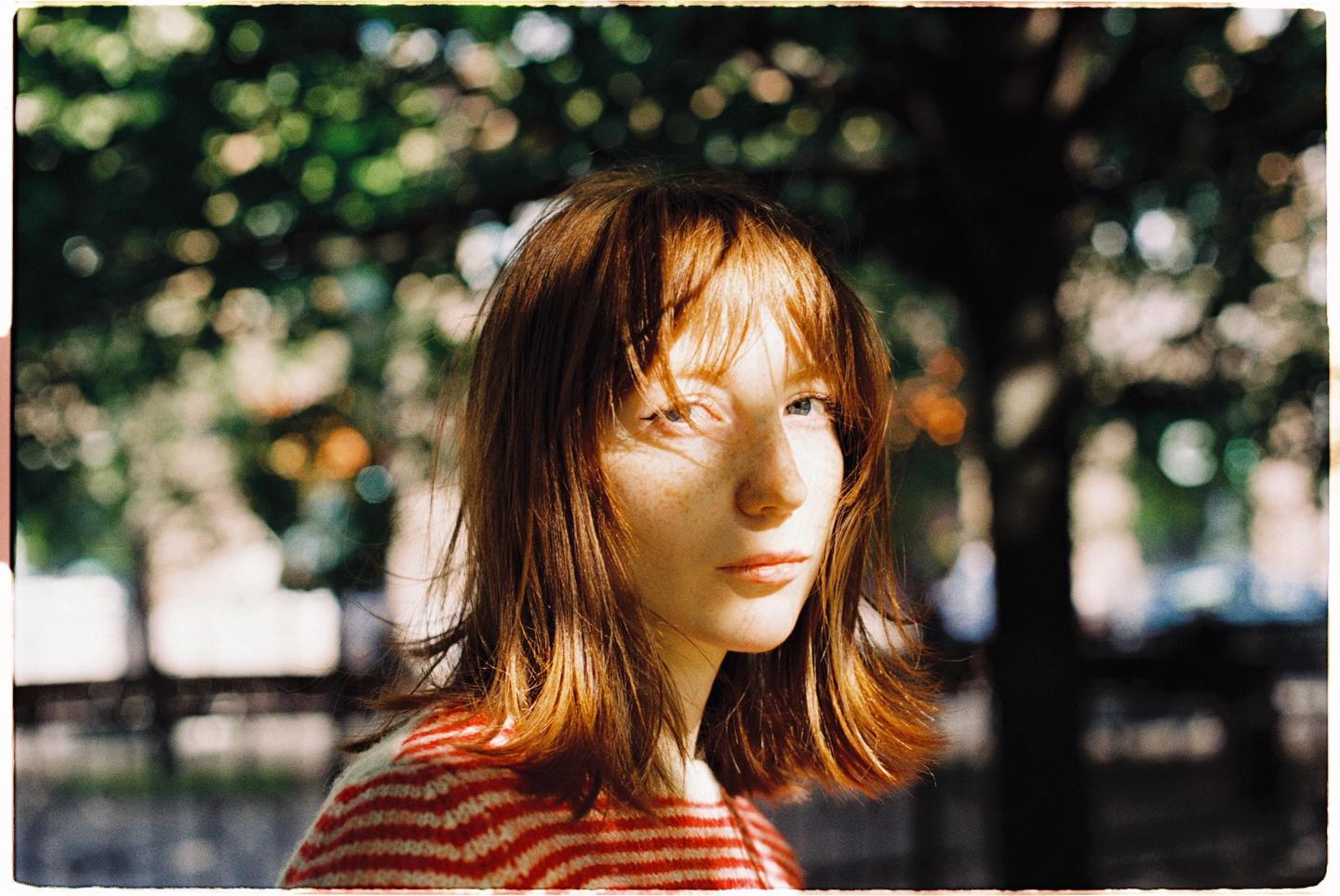 A woman with red hair and freckles looks to the side, illuminated by sunlight filtering through trees.