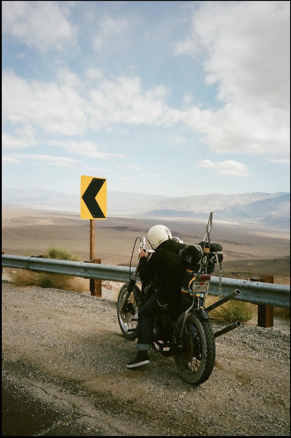 A person on a motorcycle is parked on the side of a desert road next to a yellow arrow sign, overlooking a vast valley.