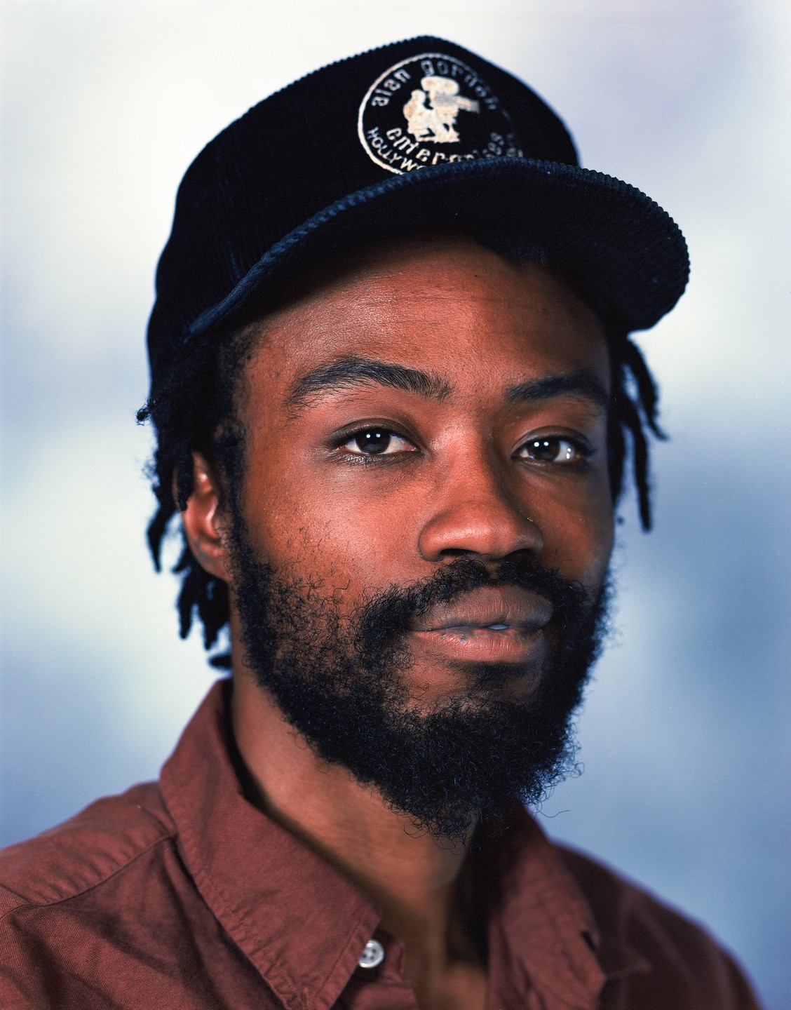 A man with a beard and dreadlocks wears a dark cap and a brown shirt.