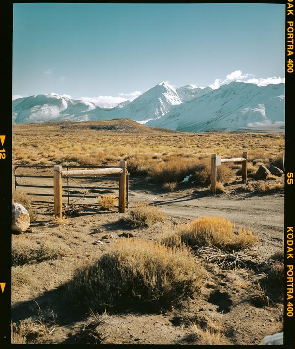A dirt road leads to a wooden gate in a dry, grassy landscape with snow-capped mountains in the distance under a clear sky.