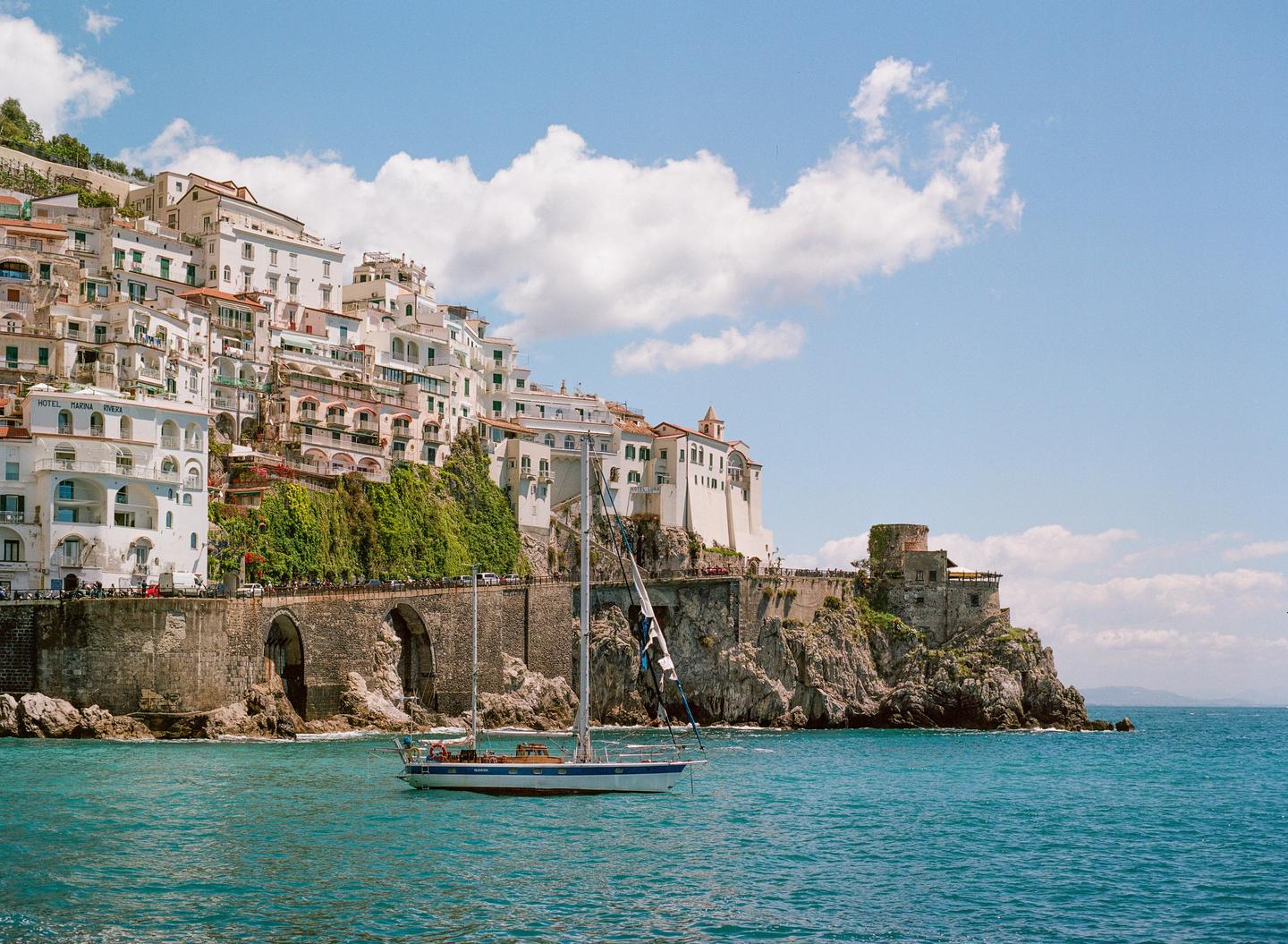 A sailboat floats on the water in front of a town built into a cliffside.