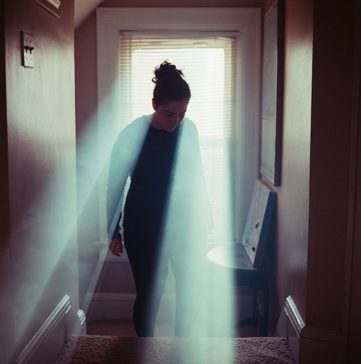 A woman stands on a carpeted staircase, illuminated by a strong beam of light from a window.