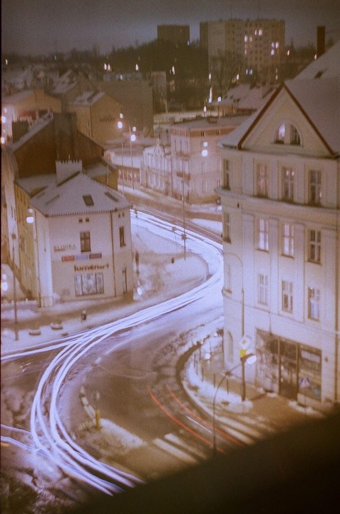A snowy street at night is illuminated by streetlights and the light trails of passing vehicles.