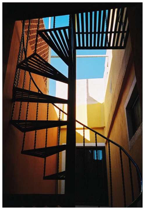 A black spiral staircase ascends between orange and yellow walls towards a bright blue sky.