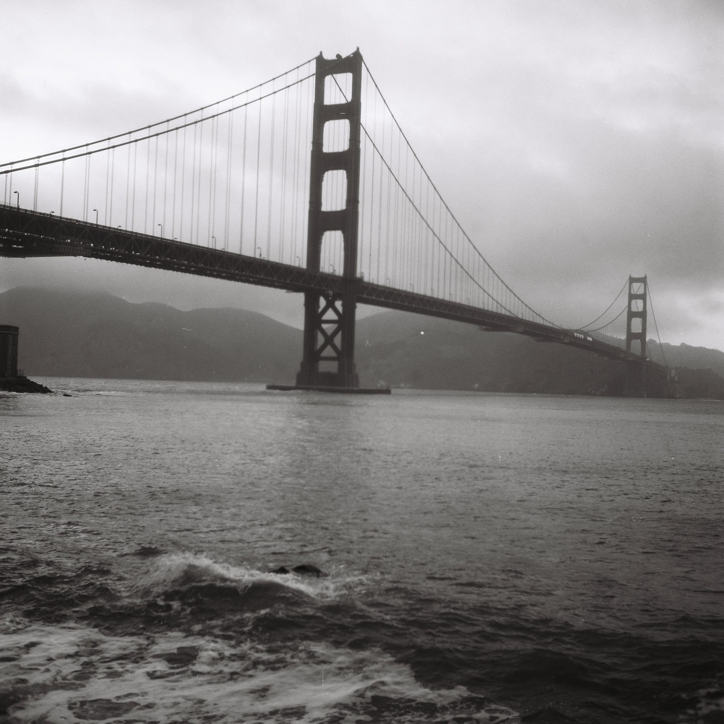 The Golden Gate Bridge spans a body of water with hills in the background under a cloudy sky.