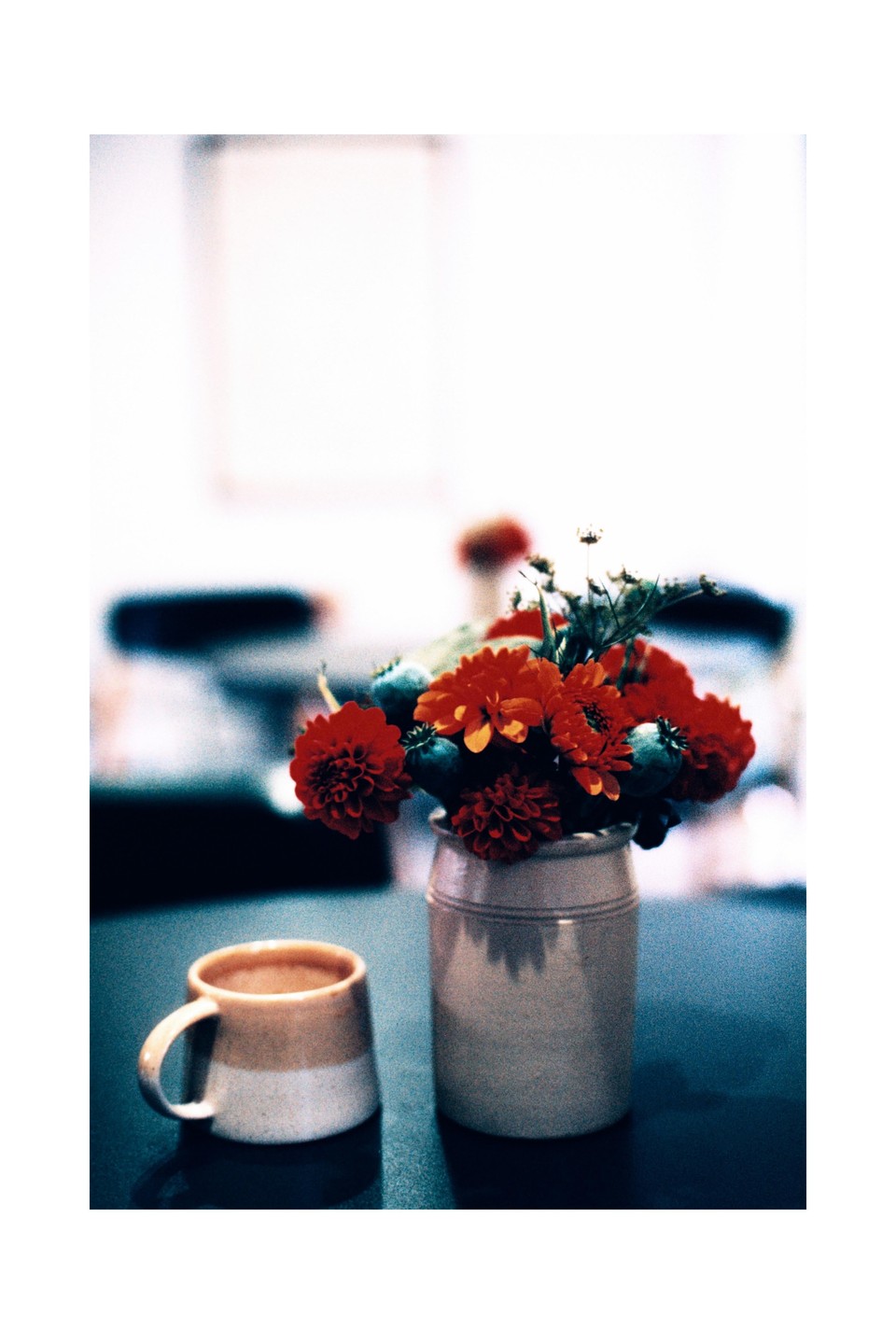A small bouquet of red flowers in a ceramic vase sits next to a two-toned mug on a dark surface.