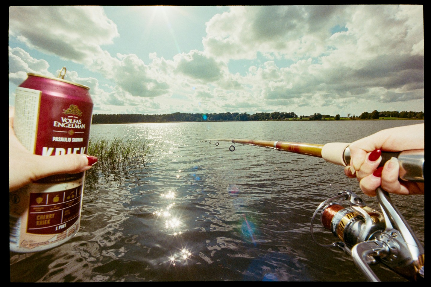 A person holds a fishing rod and a can of beer over a lake with a bright sun in the sky.