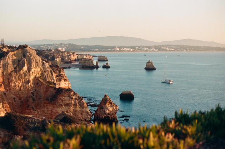 A sailboat is visible in the ocean near a rocky coastline with a distant town and mountains under a hazy sky.