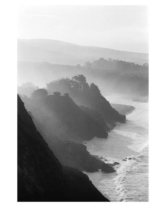 Waves crash against the rocky coastline of Big Sur, with hazy mountains and trees in the background.