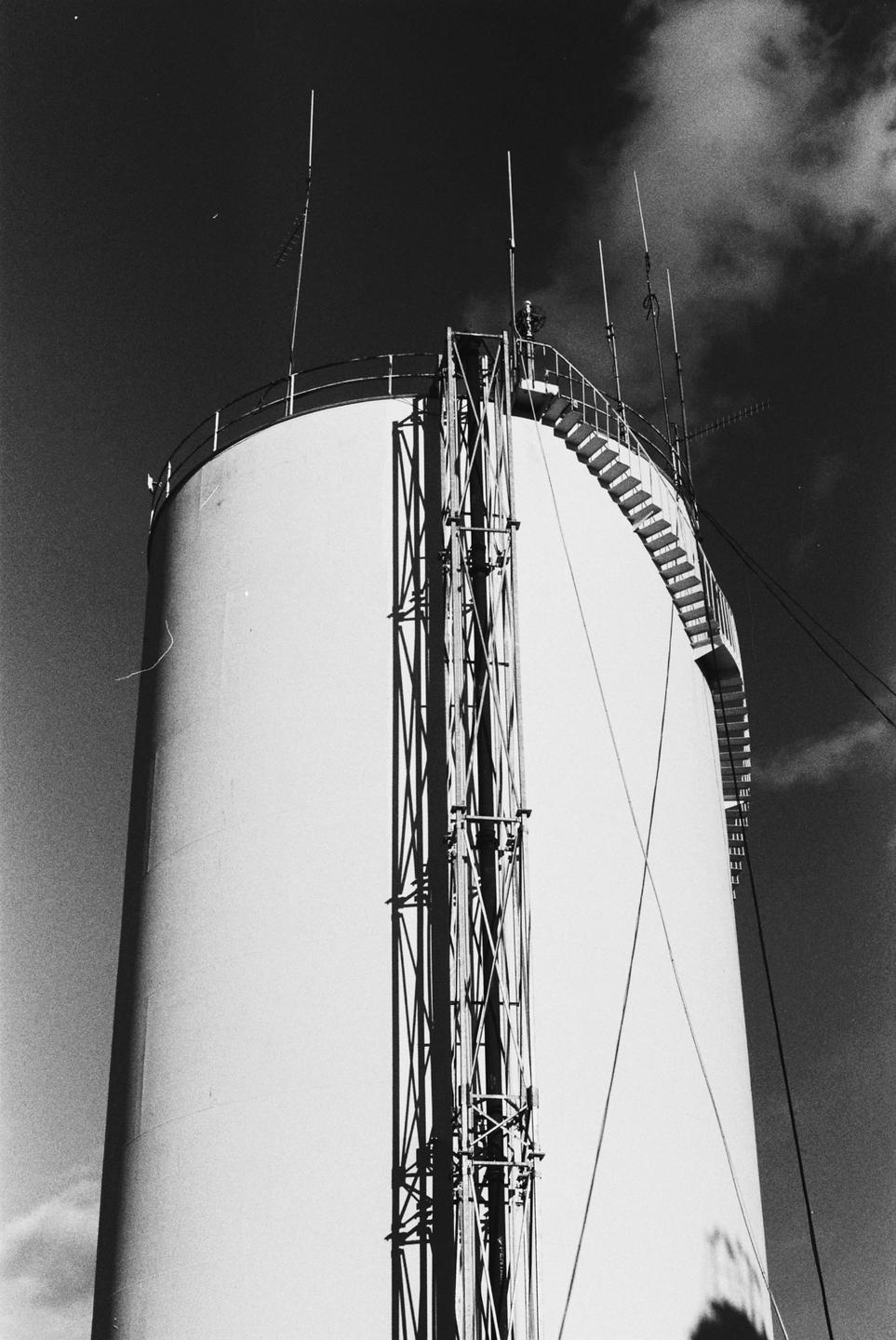 A tall, cylindrical water tower with a spiral staircase and antennas is shown against a cloudy sky.