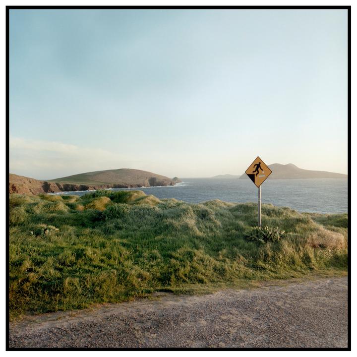 A warning sign for falling rocks stands on a grassy hill overlooking the ocean and distant landmasses.