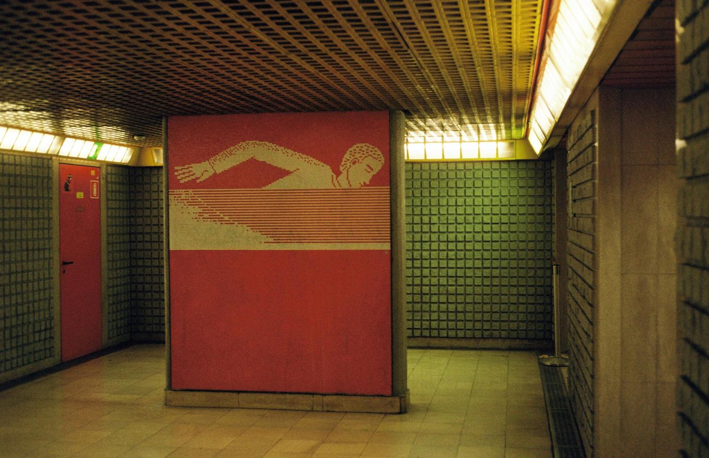 A red wall with a mosaic of a swimmer is visible in a hallway with green tiled walls and a patterned ceiling.