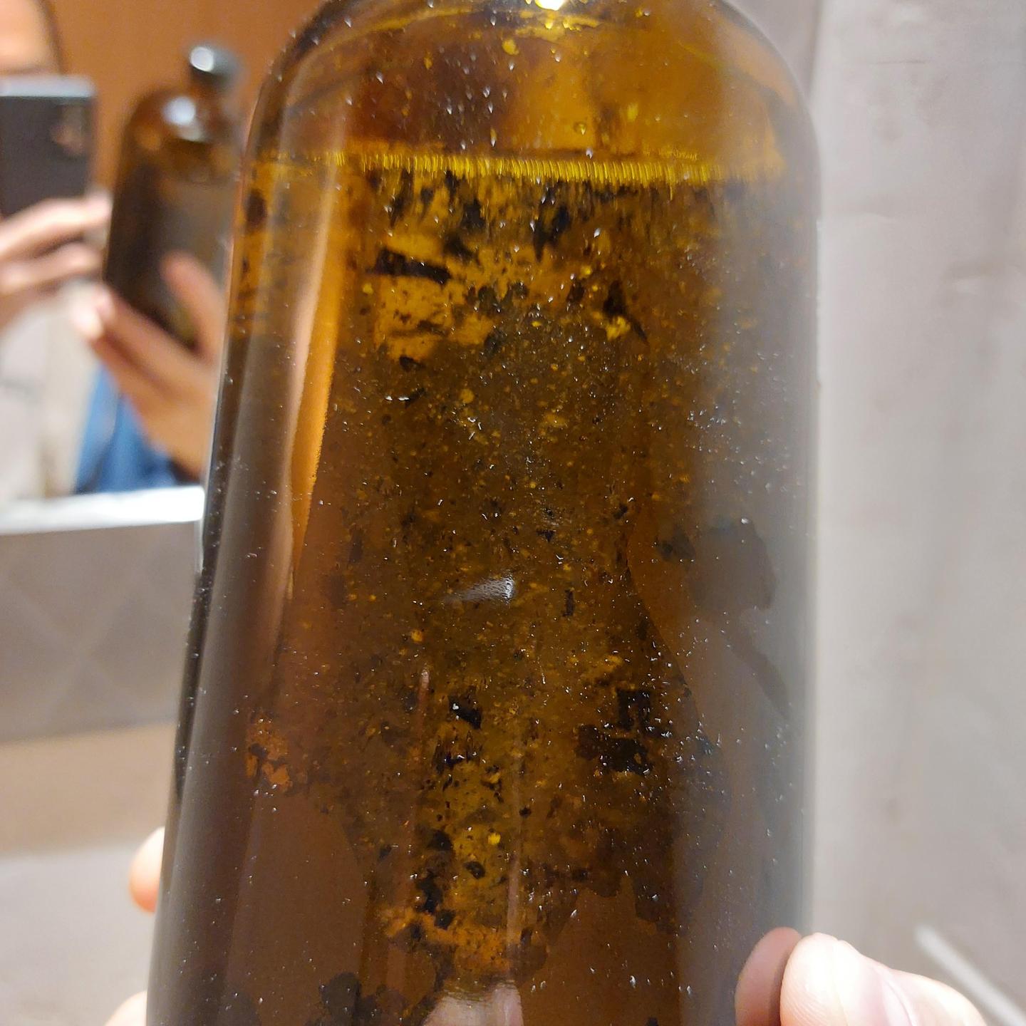 A hand holds a brown bottle filled with a dark liquid and black and yellow sediment.