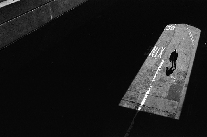 A man in a hat stands in a beam of light on a street with painted markings.