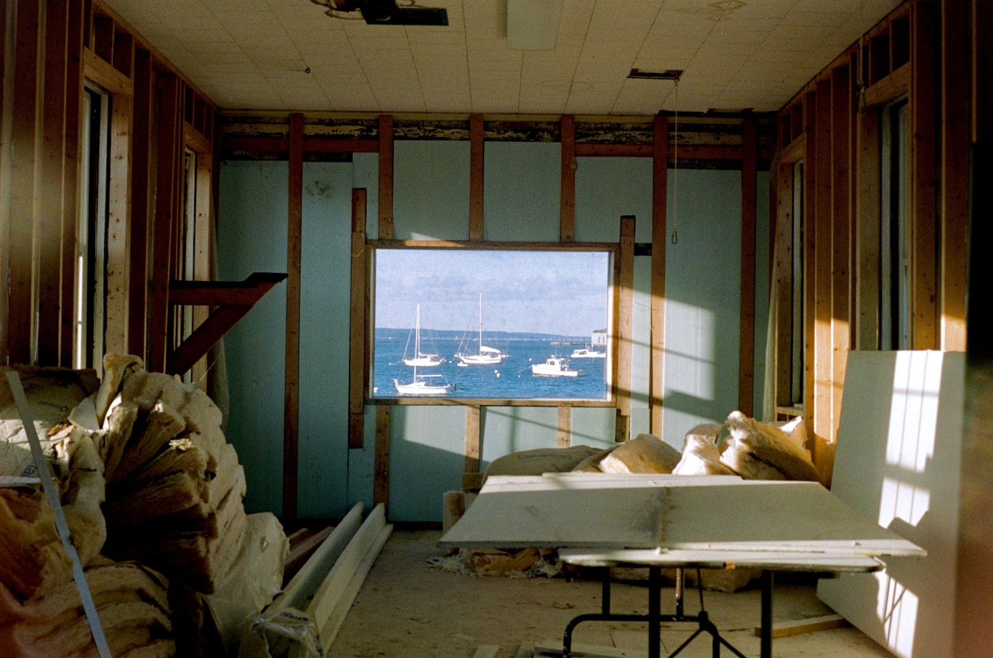 A room under construction with exposed wooden studs and insulation looks out onto a body of water with several boats.
