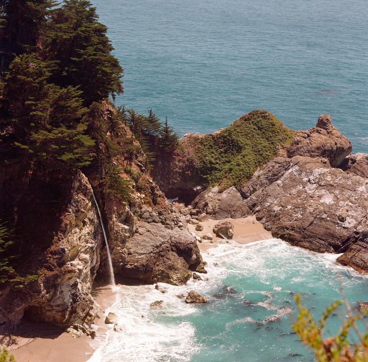 A waterfall cascades onto a sandy beach next to the ocean, surrounded by rocky cliffs and green foliage.