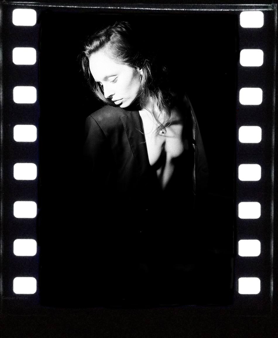A woman with long hair and a nose ring wears a suit jacket with no shirt underneath, looking down and to the right.