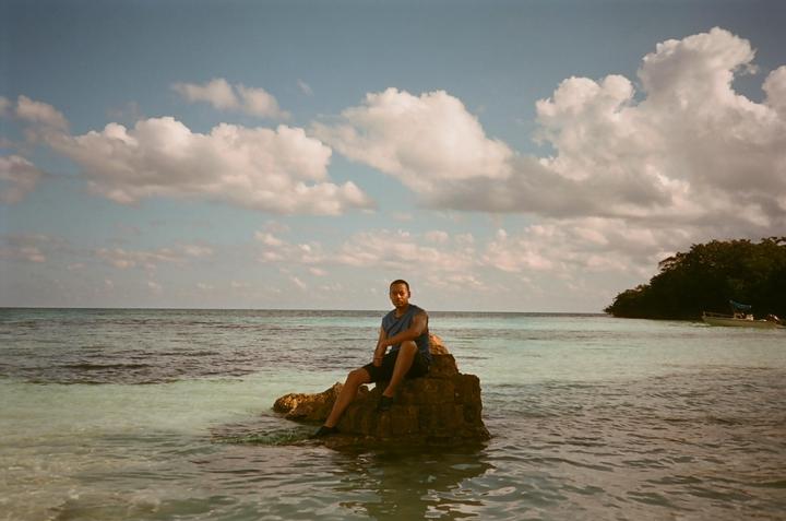 A man sits on a rock in the clear blue ocean with an island and boat in the background.